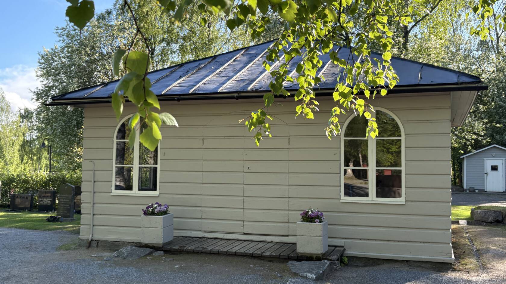 Marie kapell i vårsolen. Grå träfasad, violetta pensér i krukor vid dörren.