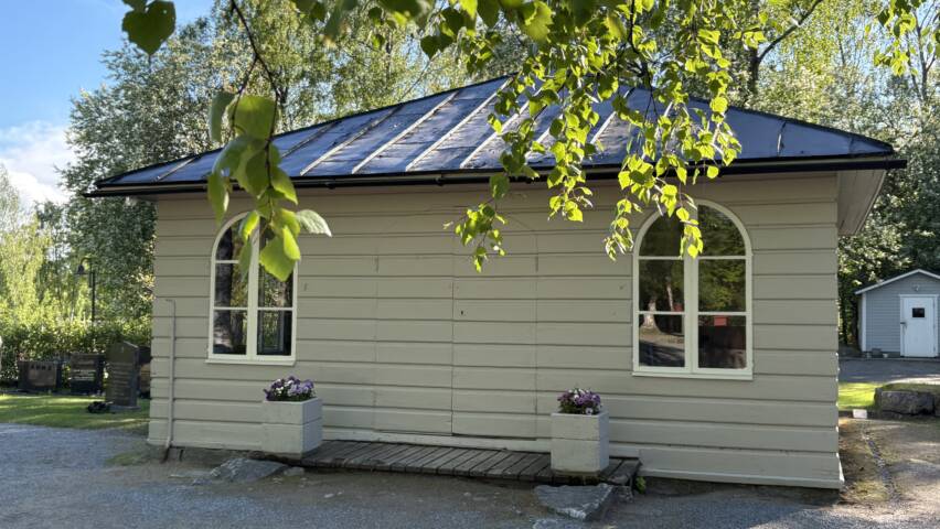 Marie kapell i vårsolen. Grå träfasad, violetta pensér i krukor vid dörren.