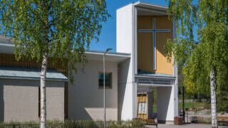 Ljus tegelfasad på Jakobstads församlingscentrum med ett vitt kors mot gul bakgrund.