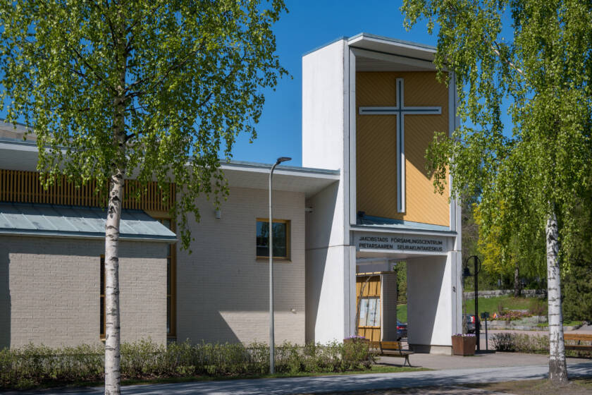 Ljus tegelfasad på Jakobstads församlingscentrum med ett vitt kors mot gul bakgrund.
