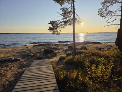 Kvällssol vid havsstrand, öppet hav. Träspång ut till klipporna i förgrunden