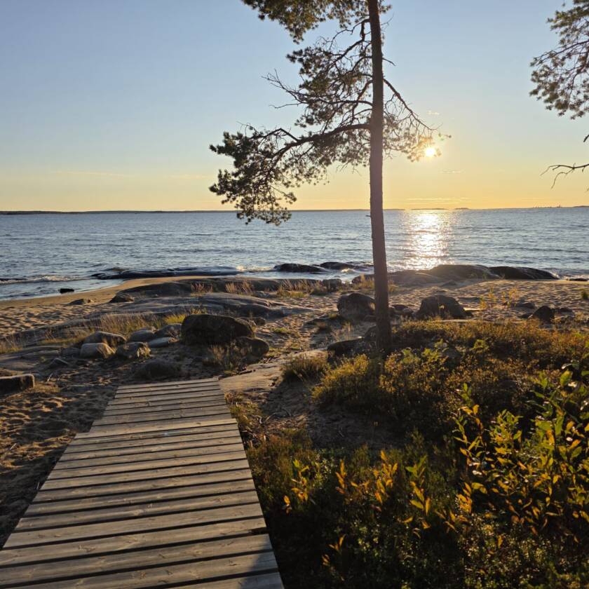 Kvällssol vid havsstrand, öppet hav. Träspång ut till klipporna i förgrunden